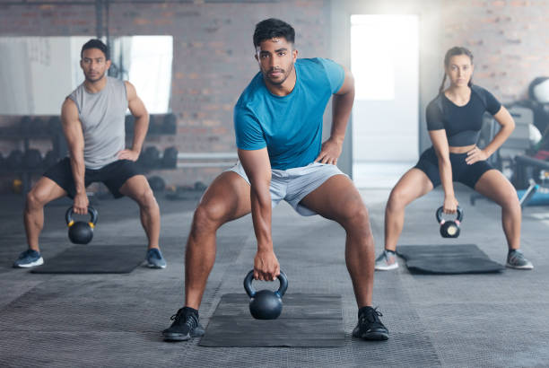 Dynamic fitness enthusiasts engage in a kettlebell workout for strength and wellness, led by a motivational personal trainer in a vibrant gym studio. Sports, motivation, and athletes performing squat exercises.