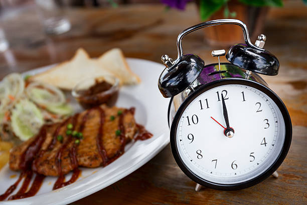 Clock on a wooden table with a steak in the background, illustrating the lunch time concept and meal timing.